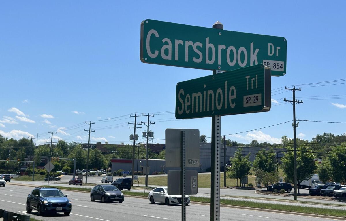 Green street signs at an intersection reading 'Carbsbrook Dr' and 'Seminole Trl' against a clear blue sky with traffic and power lines nearby.