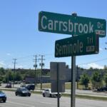 Green street signs at an intersection reading 'Carbsbrook Dr' and 'Seminole Trl' against a clear blue sky with traffic and power lines nearby.