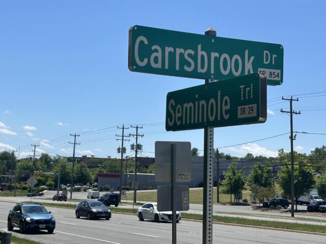 Green street signs at an intersection reading 'Carbsbrook Dr' and 'Seminole Trl' against a clear blue sky with traffic and power lines nearby.