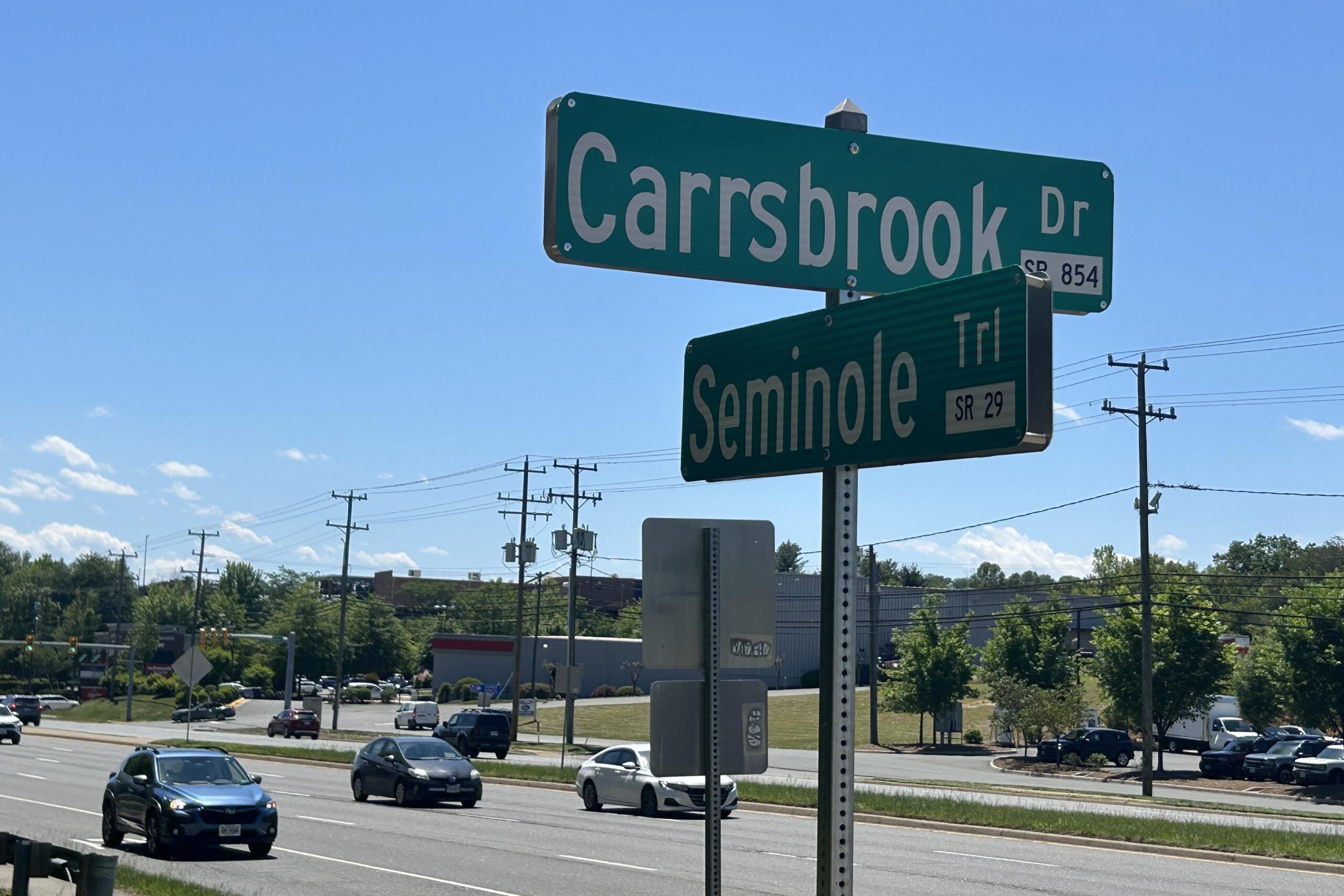 Green street signs at an intersection reading 'Carbsbrook Dr' and 'Seminole Trl' against a clear blue sky with traffic and power lines nearby.