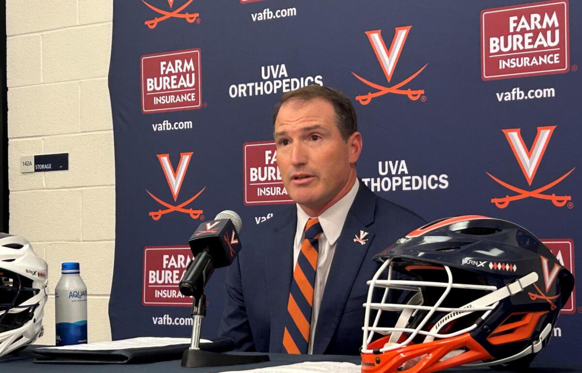 Head-and-shoulders of a man in a suit speaking at a press conference, with microphones in front and a branded Virginia backdrop behind him.