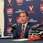 Head-and-shoulders of a man in a suit speaking at a press conference, with microphones in front and a branded Virginia backdrop behind him.