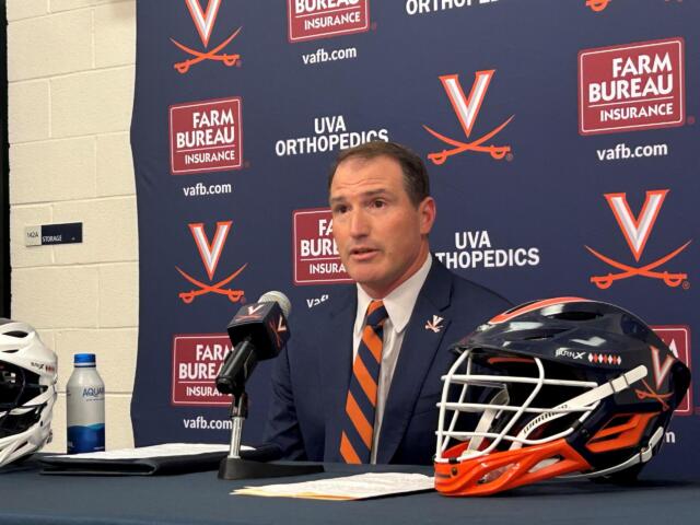 Head-and-shoulders of a man in a suit speaking at a press conference, with microphones in front and a branded Virginia backdrop behind him.