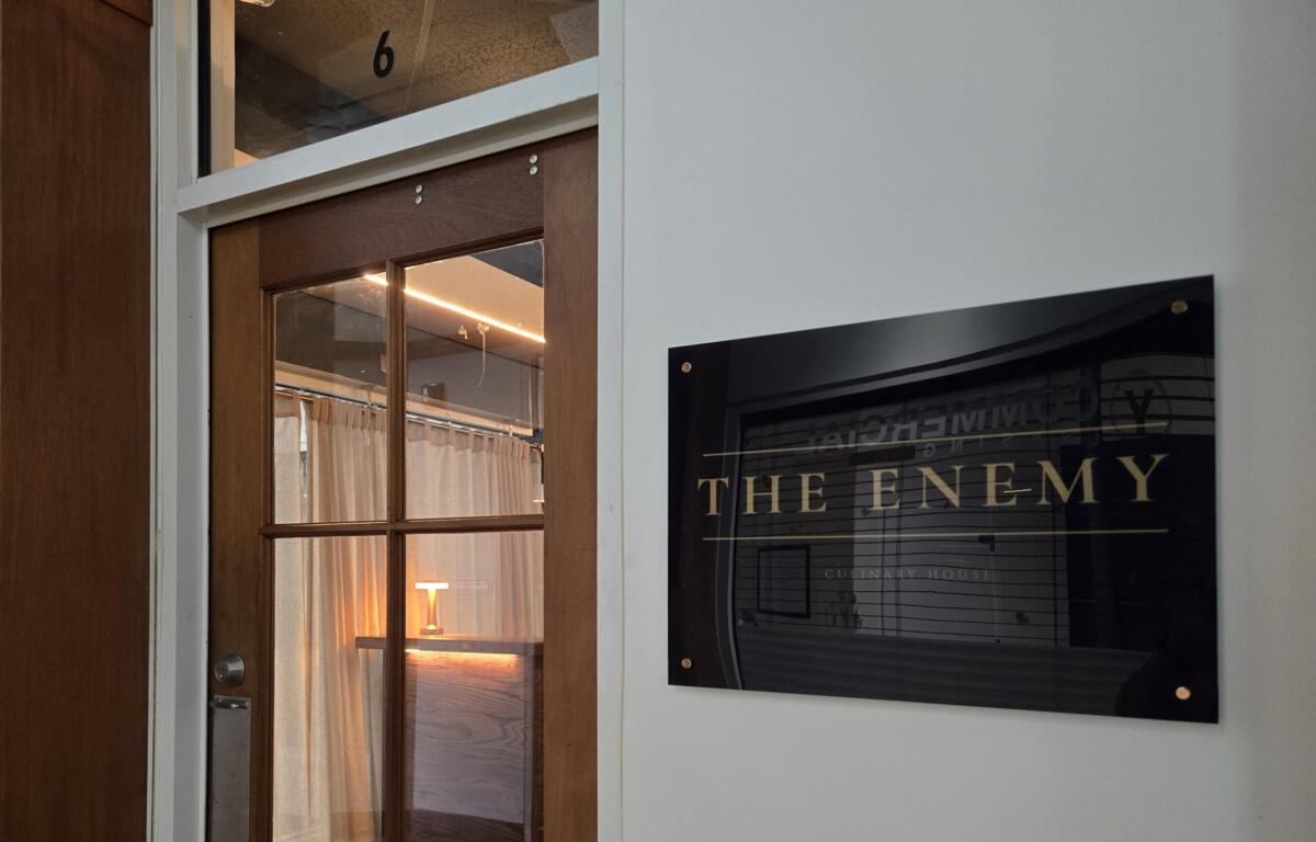 Wooden door with a glass panel next to a white wall; a poster on the wall reads 'THE ENEMY' in large letters.