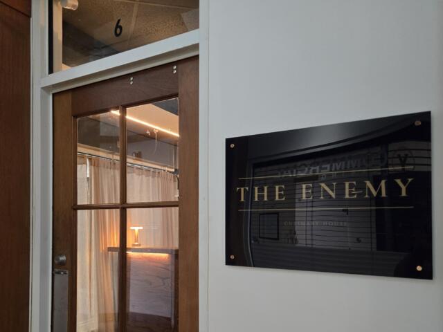 Wooden door with a glass panel next to a white wall; a poster on the wall reads 'THE ENEMY' in large letters.