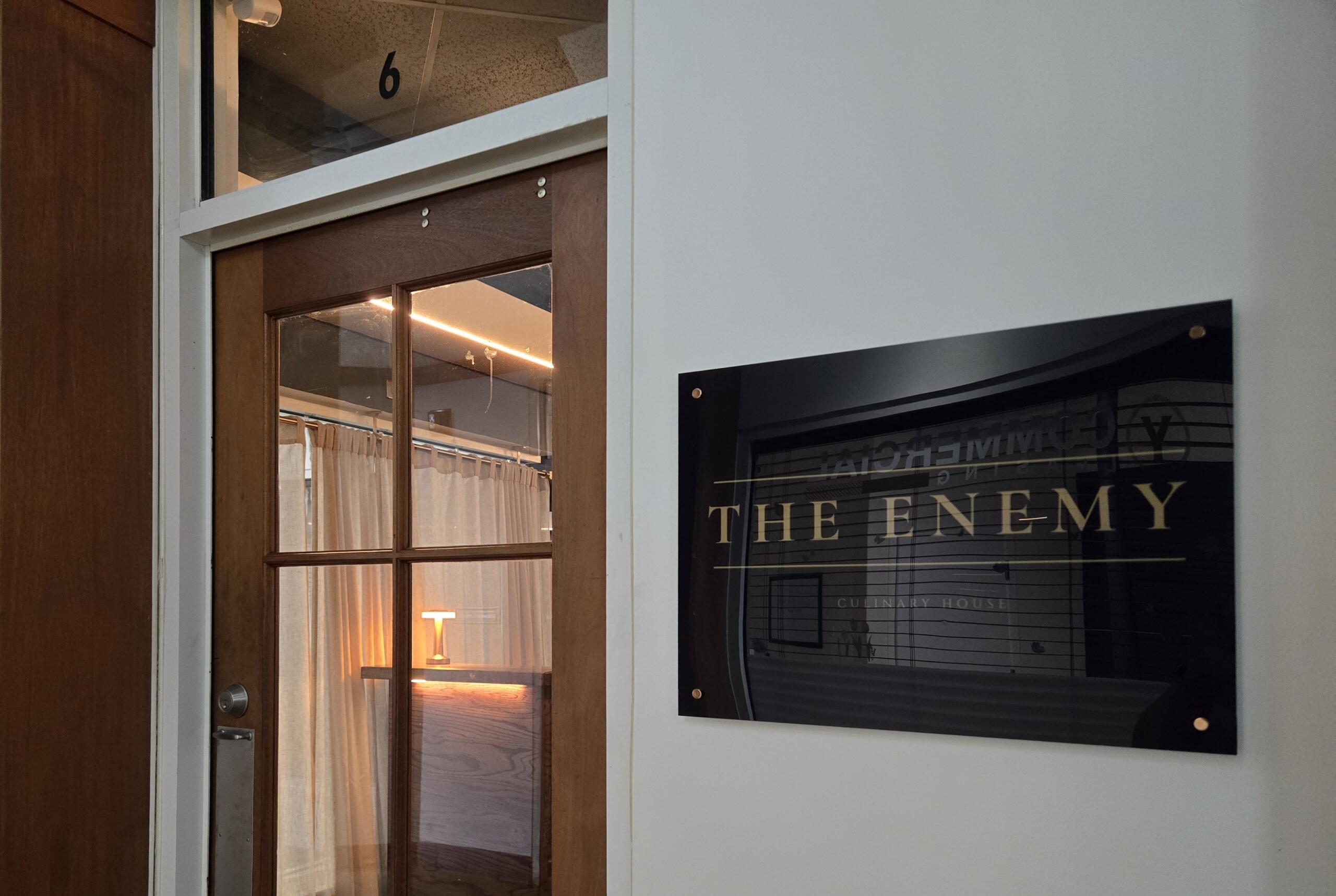 Wooden door with a glass panel next to a white wall; a poster on the wall reads 'THE ENEMY' in large letters.