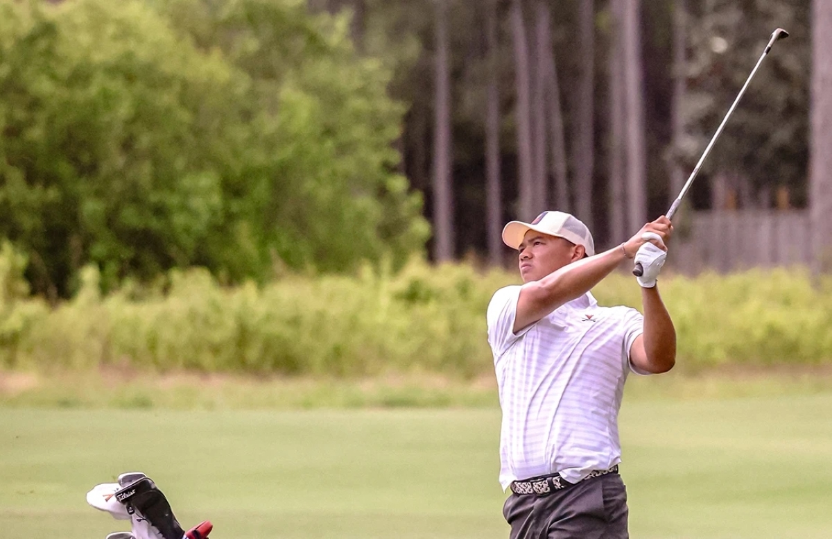 A man in a white polo shirt and cap swings a golf club on a sunny course as a bag rests nearby.