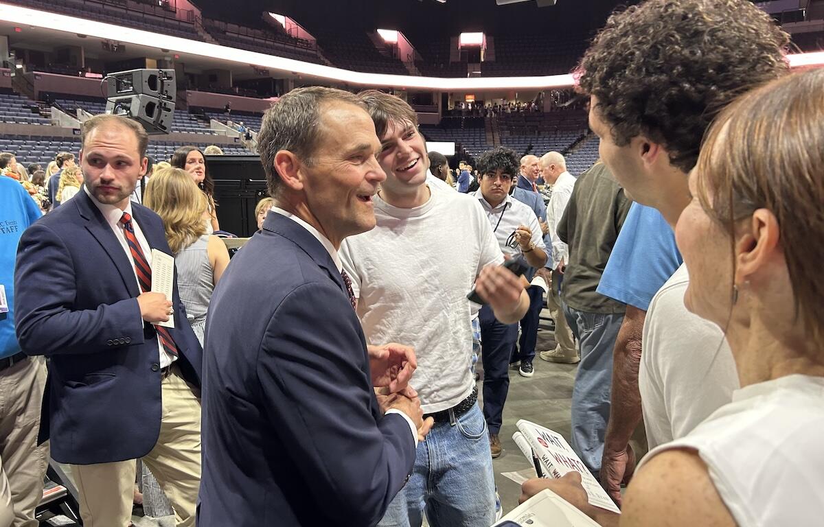 Two men shake hands and chat at a crowded event in a stadium arena, smiling and surrounded by attendees.