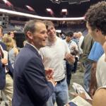 Two men shake hands and chat at a crowded event in a stadium arena, smiling and surrounded by attendees.