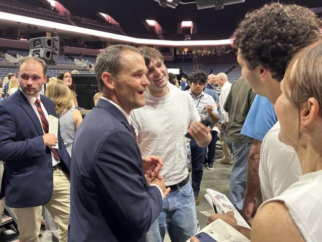 Two men shake hands and chat at a crowded event in a stadium arena, smiling and surrounded by attendees.