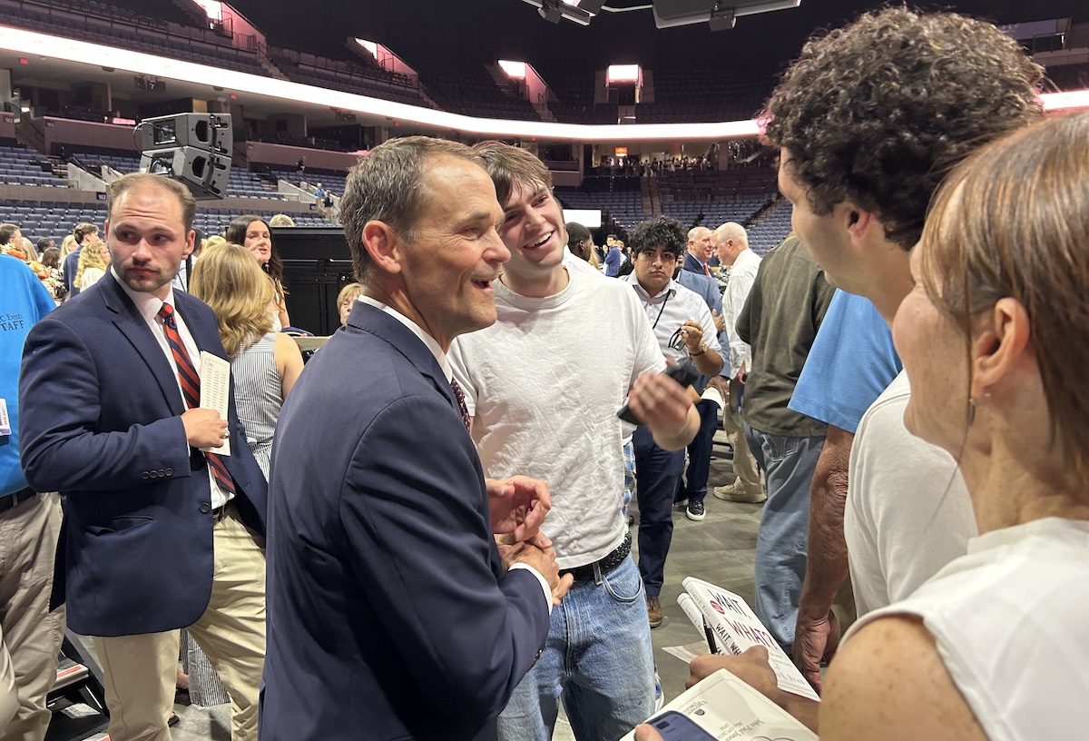 Two men shake hands and chat at a crowded event in a stadium arena, smiling and surrounded by attendees.