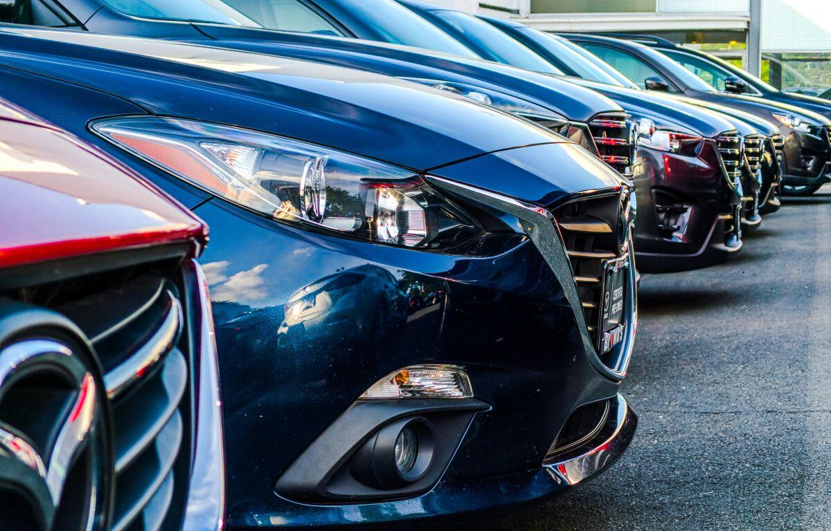 Line of new cars parked closely in a dealership lot, front ends facing the camera, blue car prominent in foreground.