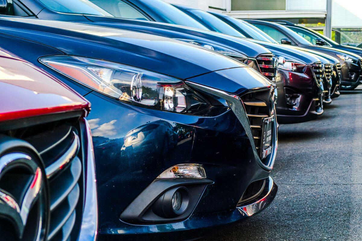 Line of new cars parked closely in a dealership lot, front ends facing the camera, blue car prominent in foreground.