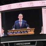 Male speaker at a wooden podium delivering a speech on a large curved LED stage with United Bank signage visible beneath.