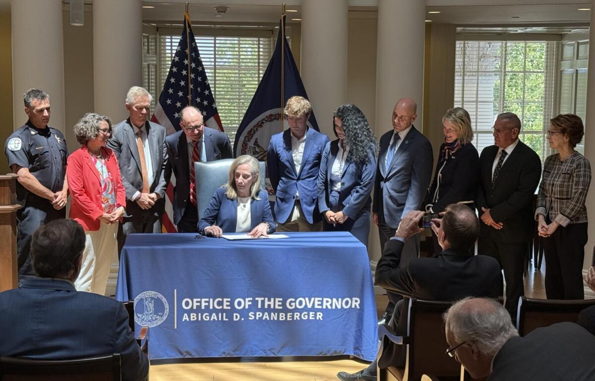 Woman in a navy blazer sits at a table signing papers as colleagues stand behind; flags and a blue banner mark an official ceremony.