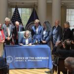 Woman in a navy blazer sits at a table signing papers as colleagues stand behind; flags and a blue banner mark an official ceremony.