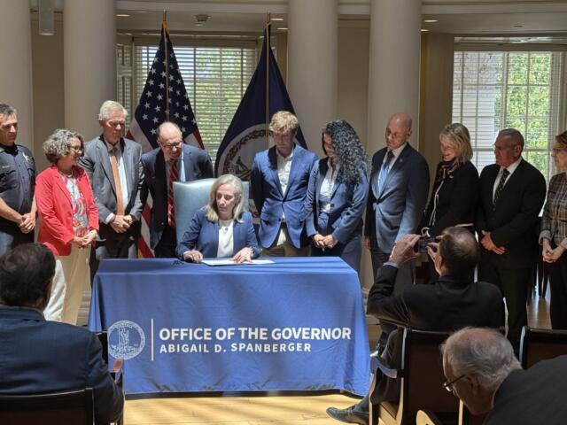 Woman in a navy blazer sits at a table signing papers as colleagues stand behind; flags and a blue banner mark an official ceremony.