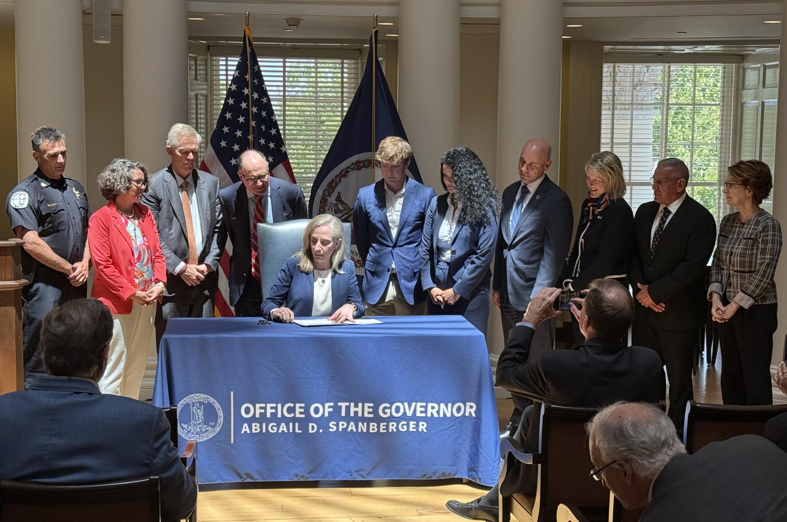 Woman in a navy blazer sits at a table signing papers as colleagues stand behind; flags and a blue banner mark an official ceremony.
