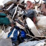 Large pile of construction debris, furniture, insulation, and a white mattress in a junkyard/landfill setting.