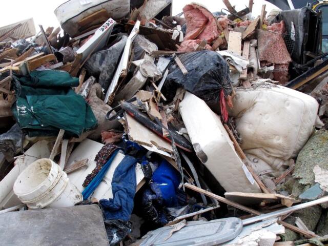 Large pile of construction debris, furniture, insulation, and a white mattress in a junkyard/landfill setting.
