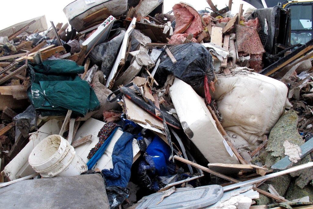 Large pile of construction debris, furniture, insulation, and a white mattress in a junkyard/landfill setting.