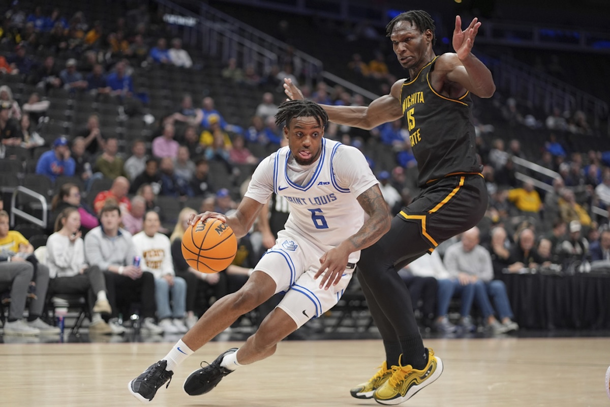 Saint Louis player #6 in white dribbles the ball while a Wichita State defender in black and gold reaches in.