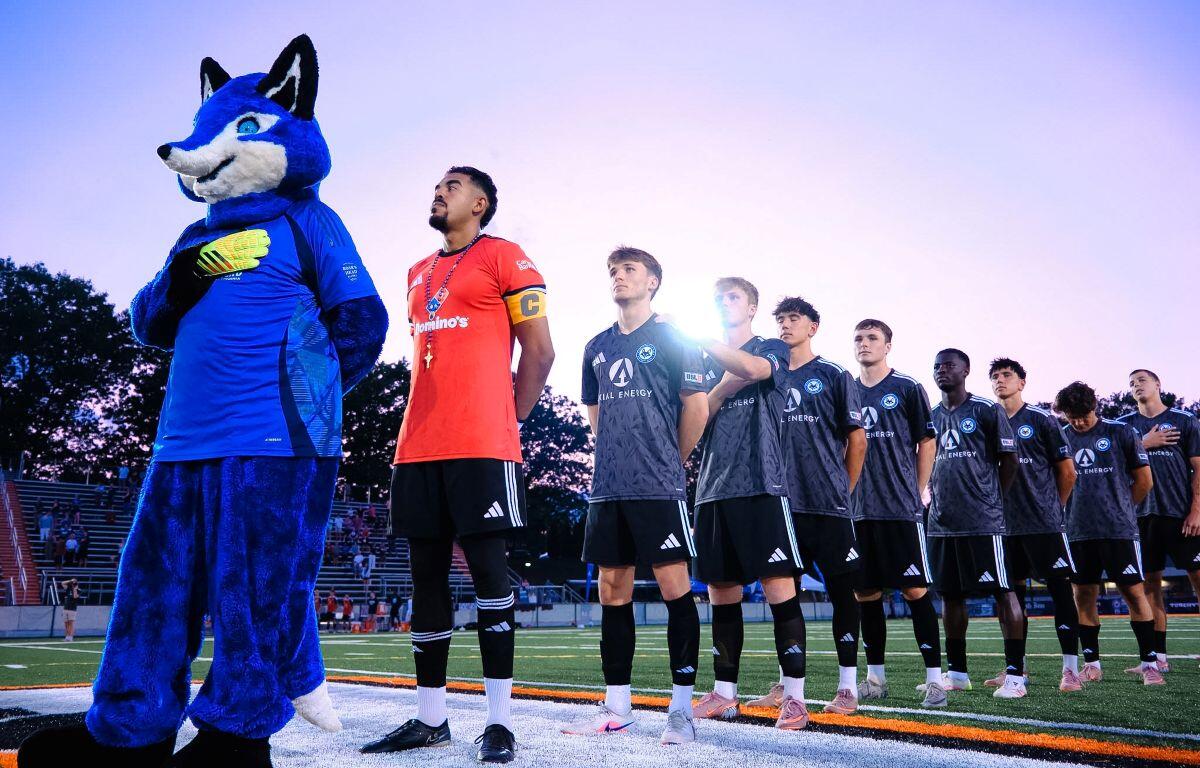 Soccer team lines up on the field with a blue fox mascot at left, before a game at sunset/blue hour.