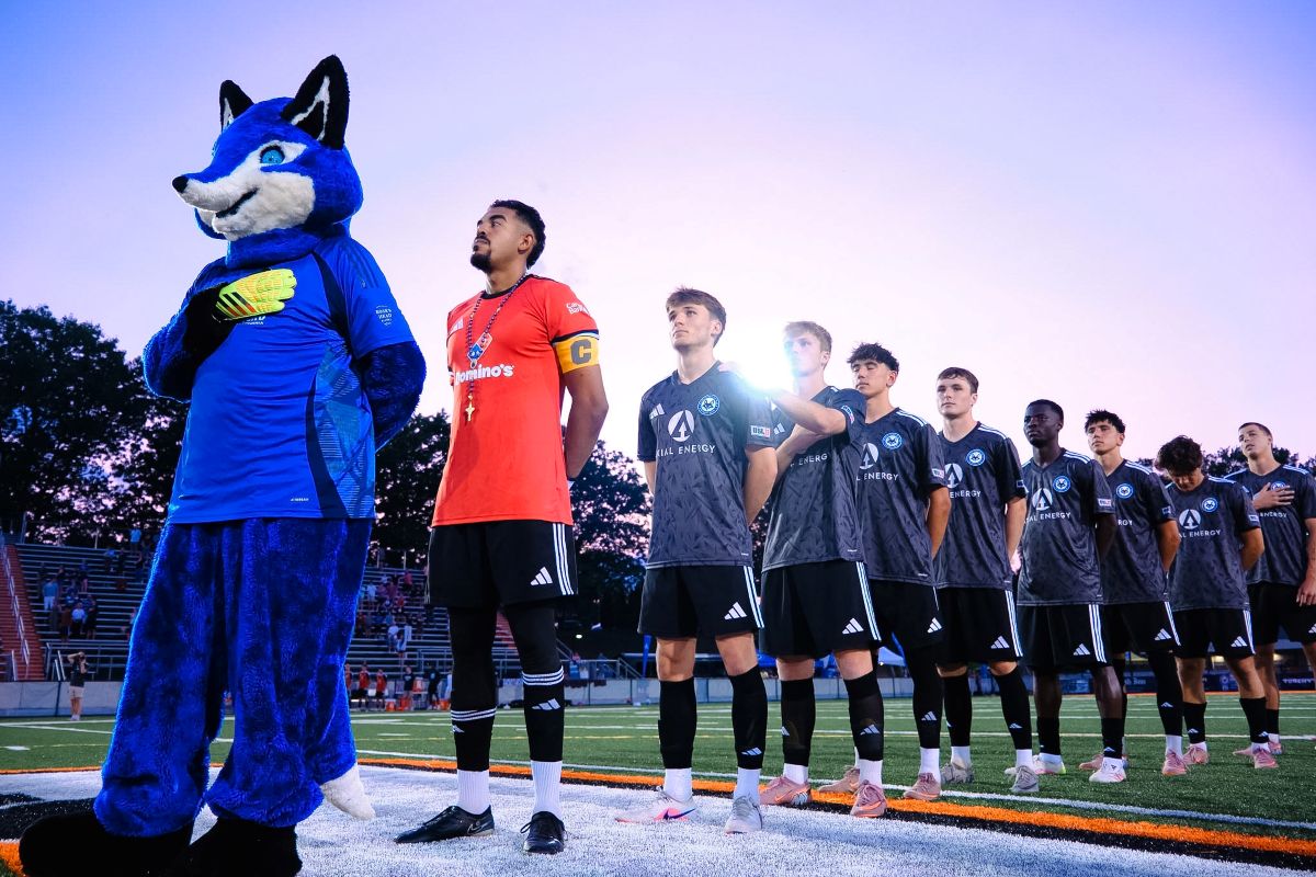 Soccer team lines up on the field with a blue fox mascot at left, before a game at sunset/blue hour.