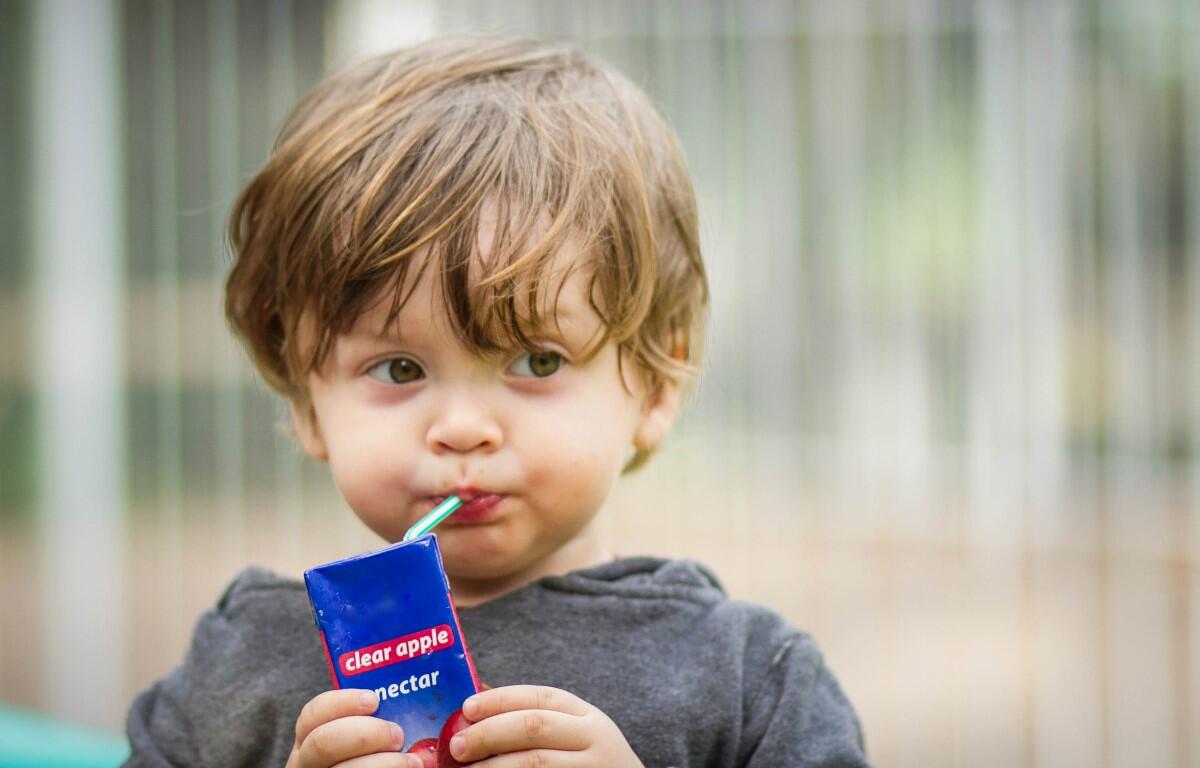 Toddler sipping juice through a green straw from a blue juice box labeled “clear apple nectar” at a playground.