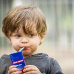 Toddler sipping juice through a green straw from a blue juice box labeled “clear apple nectar” at a playground.