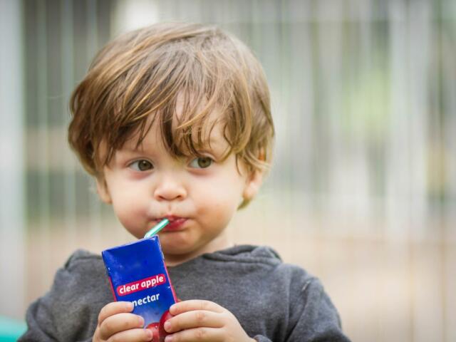 Toddler sipping juice through a green straw from a blue juice box labeled “clear apple nectar” at a playground.