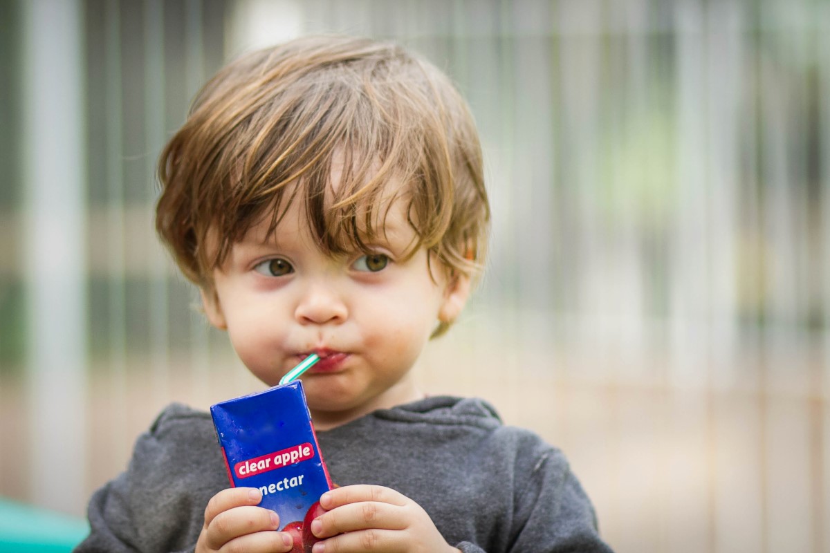 Toddler sipping juice through a green straw from a blue juice box labeled “clear apple nectar” at a playground.