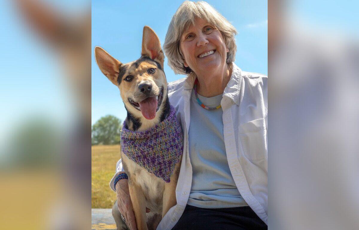 Smiling older woman sits with a happy dog wearing a purple knitted bandana on a sunny day outdoors.