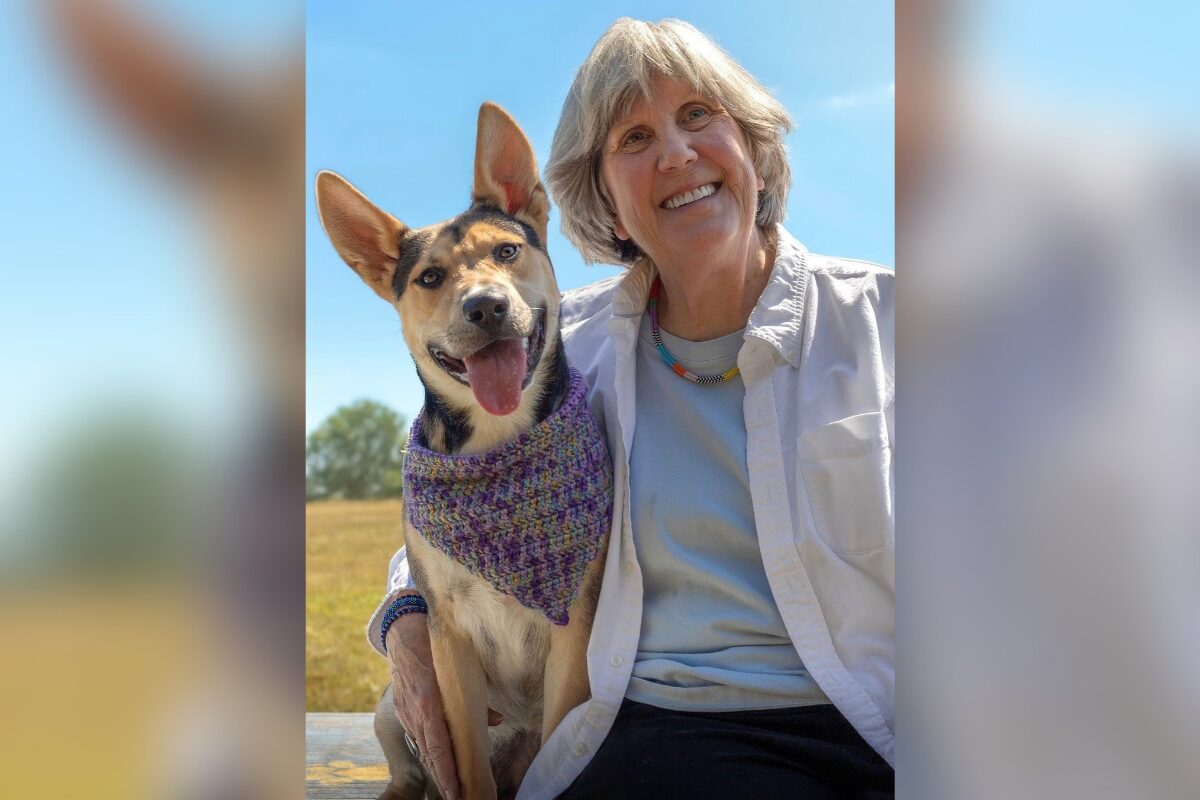 Smiling older woman sits with a happy dog wearing a purple knitted bandana on a sunny day outdoors.