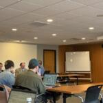 People sit around a long conference table in a meeting room, several with laptops open and papers; a whiteboard and projector screen are in the background.