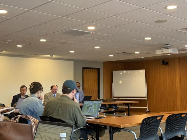 People sit around a long conference table in a meeting room, several with laptops open and papers; a whiteboard and projector screen are in the background.