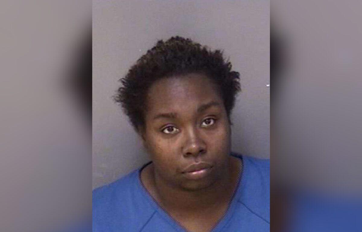 Mugshot of a young Black woman with short natural hair, wearing a blue shirt, against a gray background, looking softly at the camera.