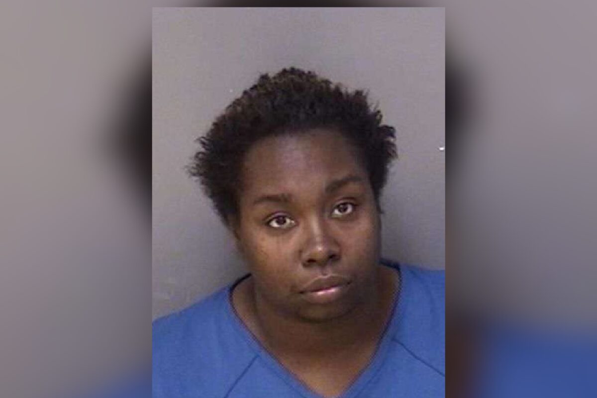 Mugshot of a young Black woman with short natural hair, wearing a blue shirt, against a gray background, looking softly at the camera.