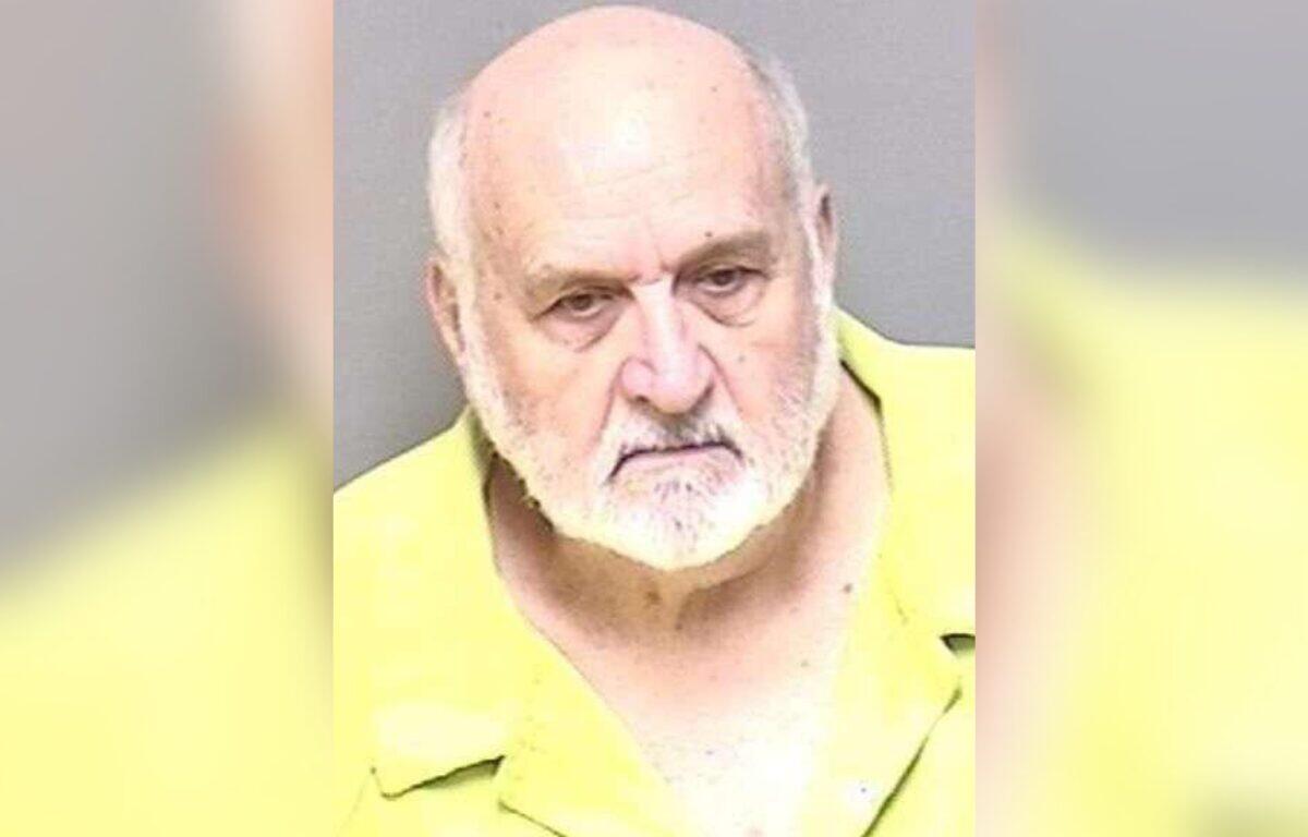 Mugshot of an elderly man with a white beard wearing a yellow prison uniform against a gray background.