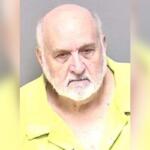 Mugshot of an elderly man with a white beard wearing a yellow prison uniform against a gray background.