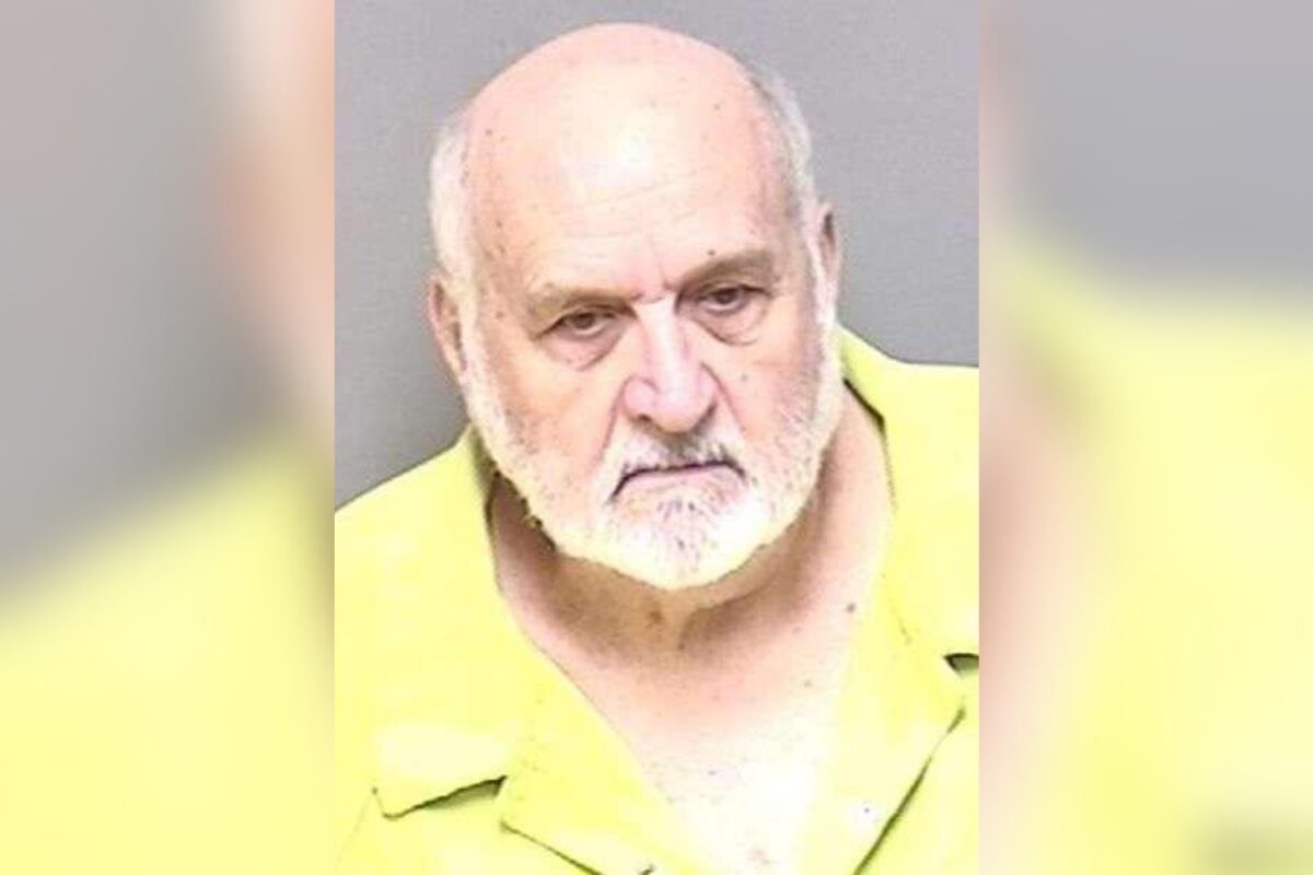 Mugshot of an elderly man with a white beard wearing a yellow prison uniform against a gray background.