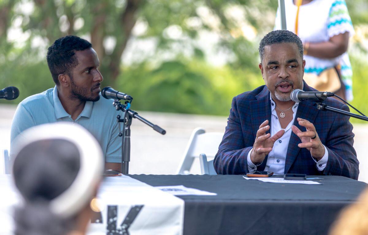 Two men sit at a panel outdoors, one speaking into a microphone while gesturing with his hands, the other listening attentively.