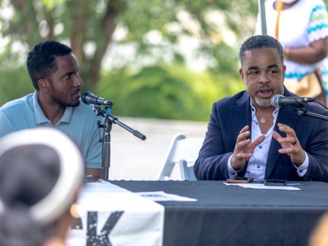 Two men sit at a panel outdoors, one speaking into a microphone while gesturing with his hands, the other listening attentively.