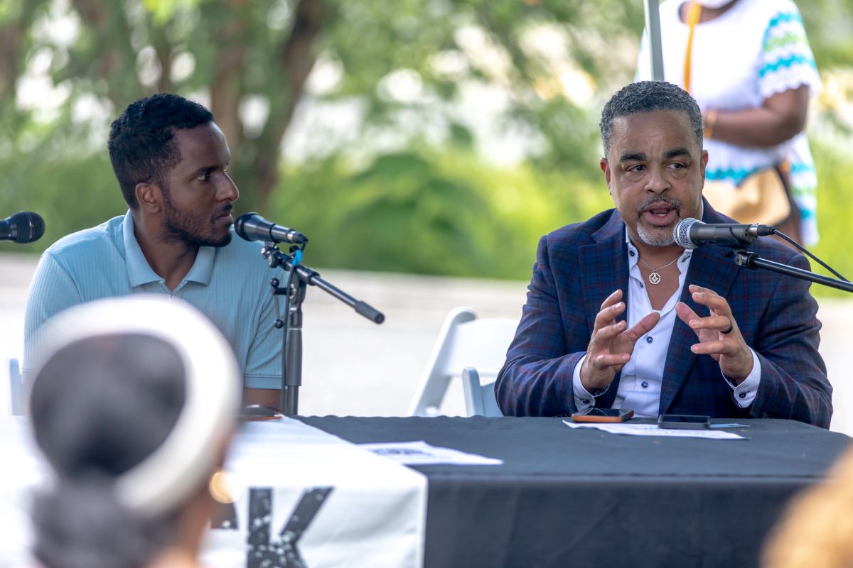 Two men sit at a panel outdoors, one speaking into a microphone while gesturing with his hands, the other listening attentively.