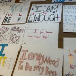 Collection of handmade posters with positive affirmations like 'I am enough' and 'Love yourself' displayed on a table surface, colorful markers and drawings visible.