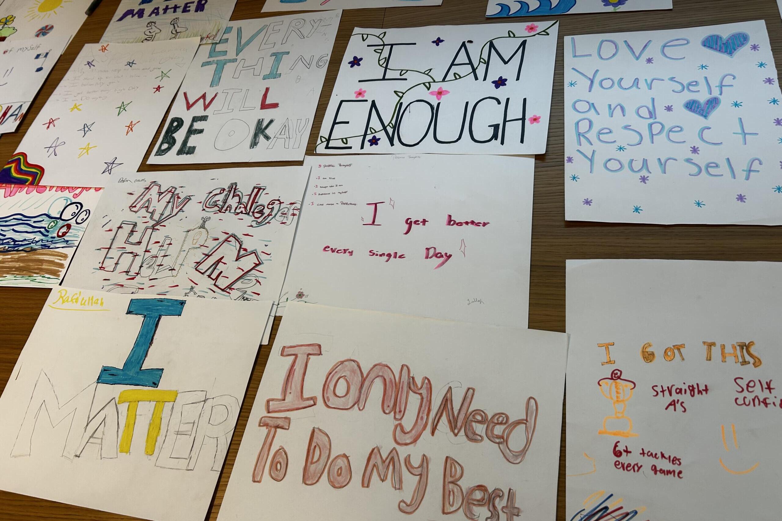 Collection of handmade posters with positive affirmations like 'I am enough' and 'Love yourself' displayed on a table surface, colorful markers and drawings visible.
