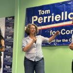 Three people on a campaign stage: a woman with a microphone speaking to a man, while another woman holds a cup, in front of a blue Tom Perrelli for Congress banner.
