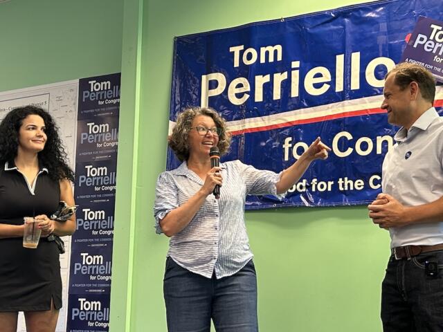 Three people on a campaign stage: a woman with a microphone speaking to a man, while another woman holds a cup, in front of a blue Tom Perrelli for Congress banner.
