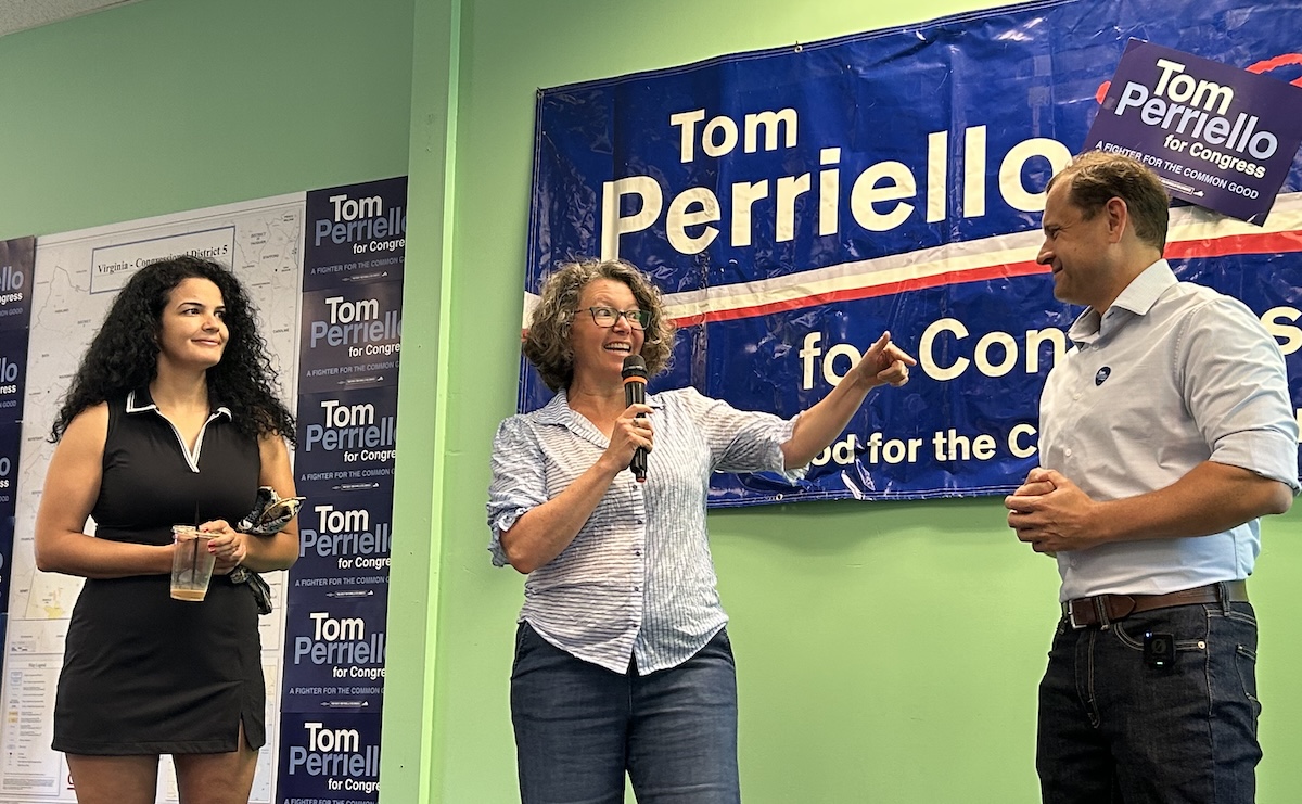 Three people on a campaign stage: a woman with a microphone speaking to a man, while another woman holds a cup, in front of a blue Tom Perrelli for Congress banner.