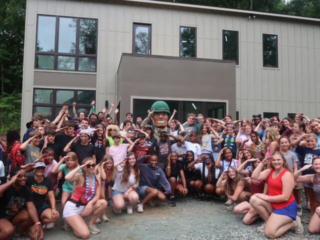 Large group of children posing outdoors in front of a modern building, with a mascot in a helmet at center, cheering and smiling.